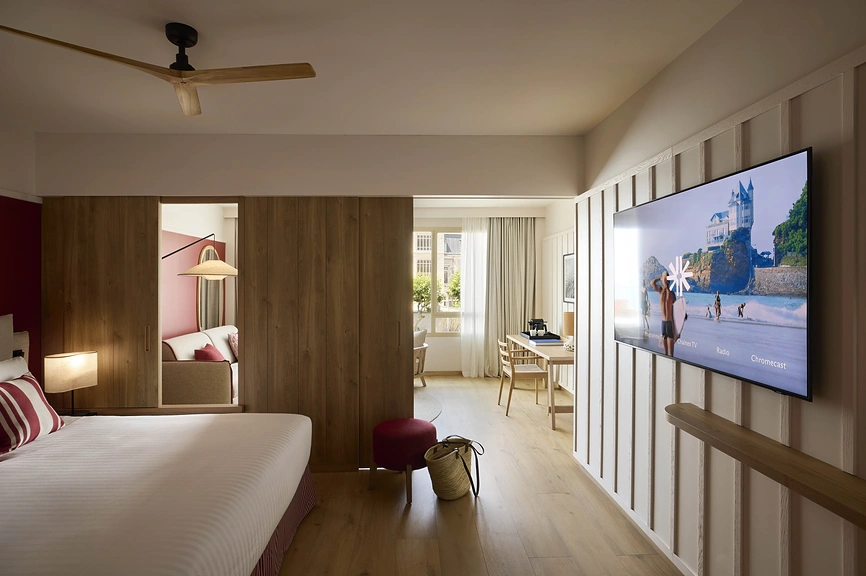 Suite hôtelière avec cloison en bois séparant chambre et espace bureau, télévision murale grand écran et ventilateur de plafond.
