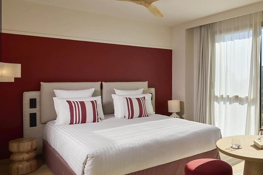 Grand lit double avec linge blanc et coussins rayés bordeaux, tête de lit en lin sur mur rouge, ventilateur de plafond et baie vitrée lumineuse.