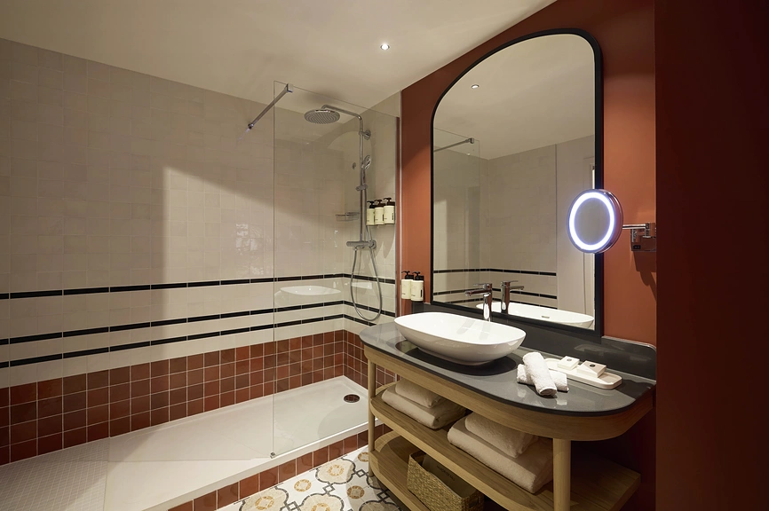 Salle de bain d'hôtel avec baignoire-douche à carrelage terracotta et blanc, vasque à poser sur meuble en chêne, grand miroir arqué et miroir de courtoisie LED.