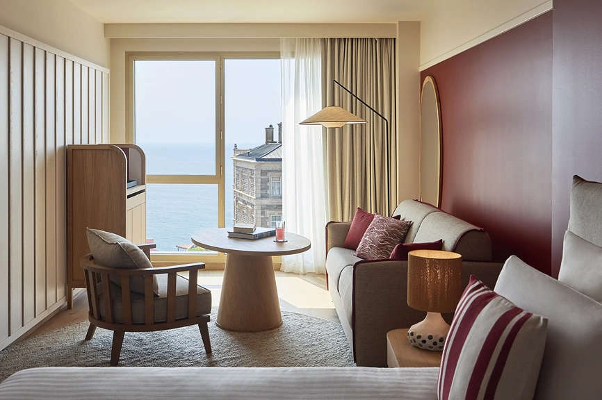 Chambre d'hôtel avec coin salon canapé et fauteuil en tons beige et bordeaux, grande fenêtre vue mer et meuble TV en chêne.