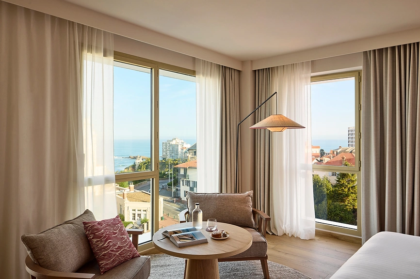 Coin salon d'une chambre d'hôtel avec fauteuil en tissu taupe, table basse ronde en bois, lampe arceau en rotin et grandes fenêtres offrant une vue panoramique sur les toits, le littoral et l'océan de Biarritz.