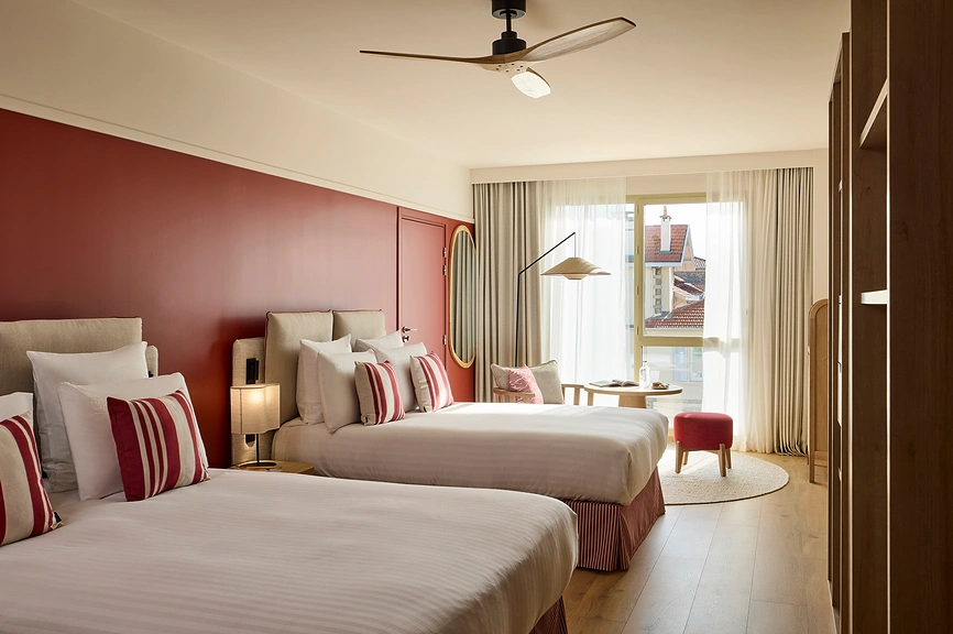 Chambre d'hôtel twin avec deux lits aux coussins rayés rouge et blanc, mur d'accent rouge bordeaux, miroir ovale doré, pouf rouge, coin salon avec table ronde et grande baie vitrée lumineuse donnant sur les toits.