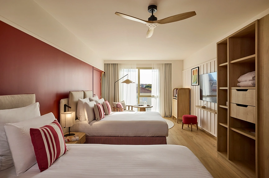 Chambre d'hôtel avec deux lits, coussins rayés rouge et blanc, mur d'accent rouge bordeaux, ventilateur de plafond, bibliothèque ouverte en bois clair avec télévision, pouf rouge et grande fenêtre lumineuse donnant sur l'extérieur.