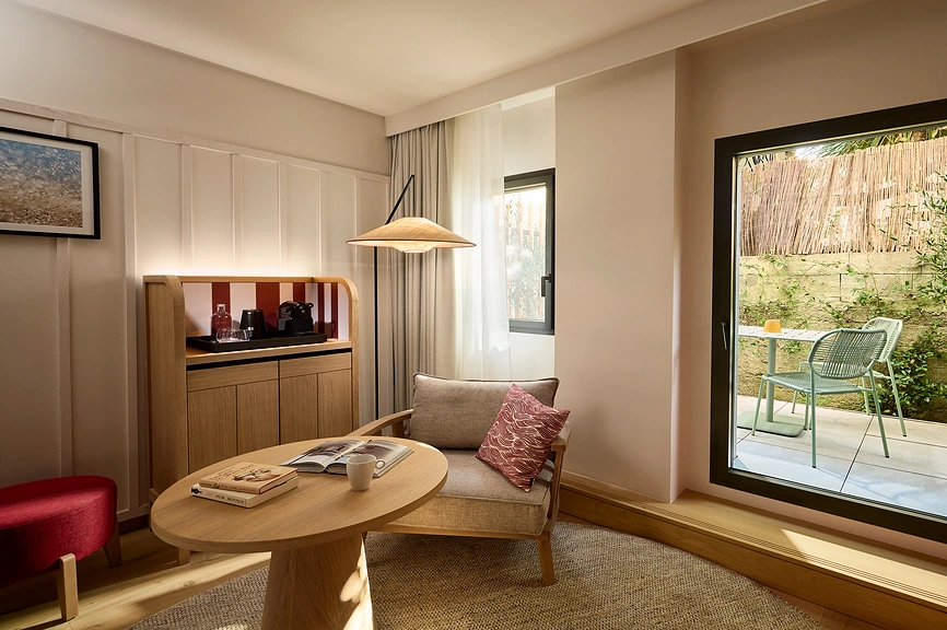 Salon d'une chambre d'hôtel lumineux avec fauteuil en tissu gris, table basse ronde en bois clair, buffet en bois avec machine à café, lampe suspendue en rotin et accès direct sur une terrasse privée avec mobilier de jardin vert sauge.