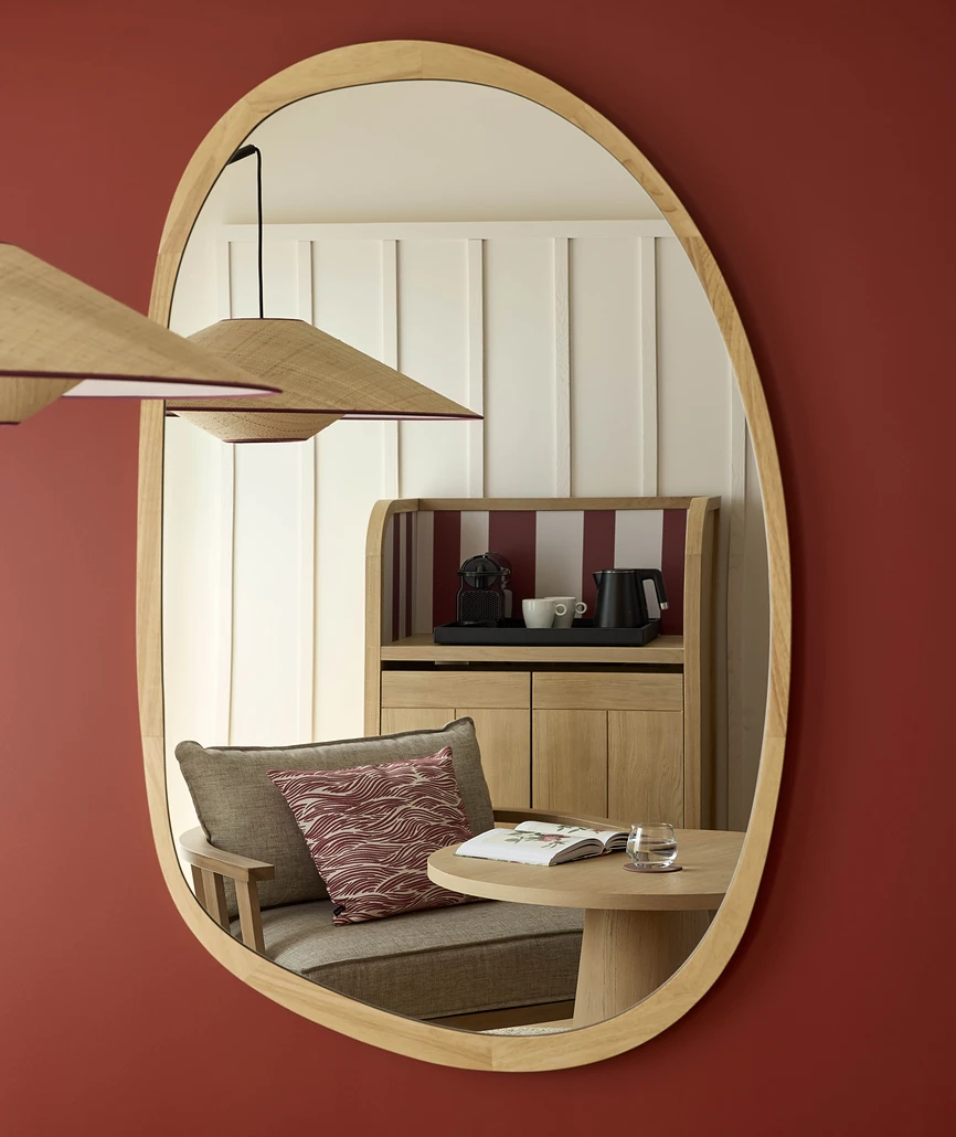 Miroir ovale en bois clair sur mur bordeaux reflétant le coin salon d'une chambre d'hôtel avec fauteuil, table basse et meuble à café en chêne.