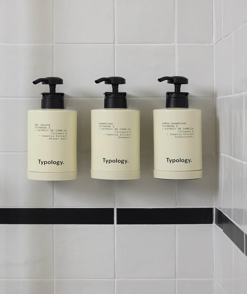 Trois flacons pompe Typology (gel douche, shampoing et après-shampoing) alignés sur un carrelage blanc en douche.