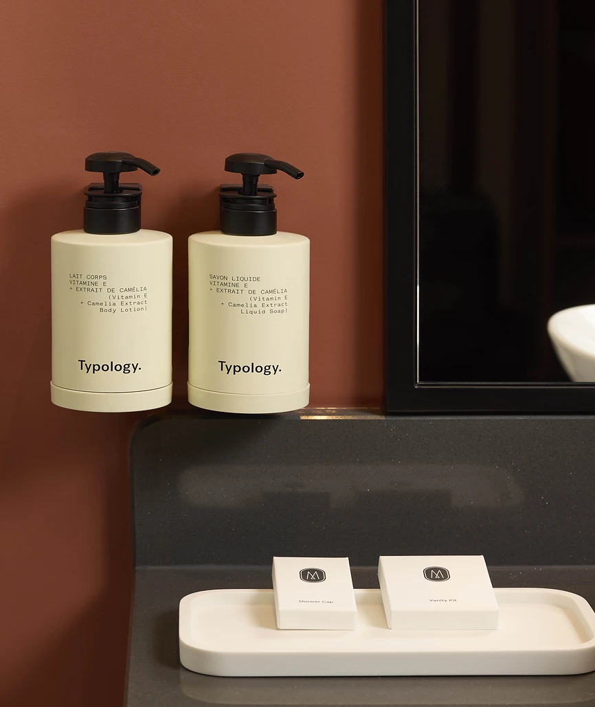 Deux flacons pompe blancs de la marque Typology (lait corps et savon liquide à la vitamine E et à l'extrait de camélia) fixés sur un mur terracotta, au-dessus d'un plan vasque en pierre noire avec des petits accessoires de salle de bain.