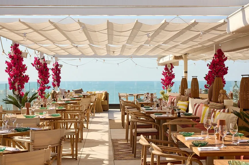 Terrasse de restaurant en rooftop sous voiles blanches, ornée de bouganvilliers fuchsia, tables en bois dressées et vue directe sur l'océan.