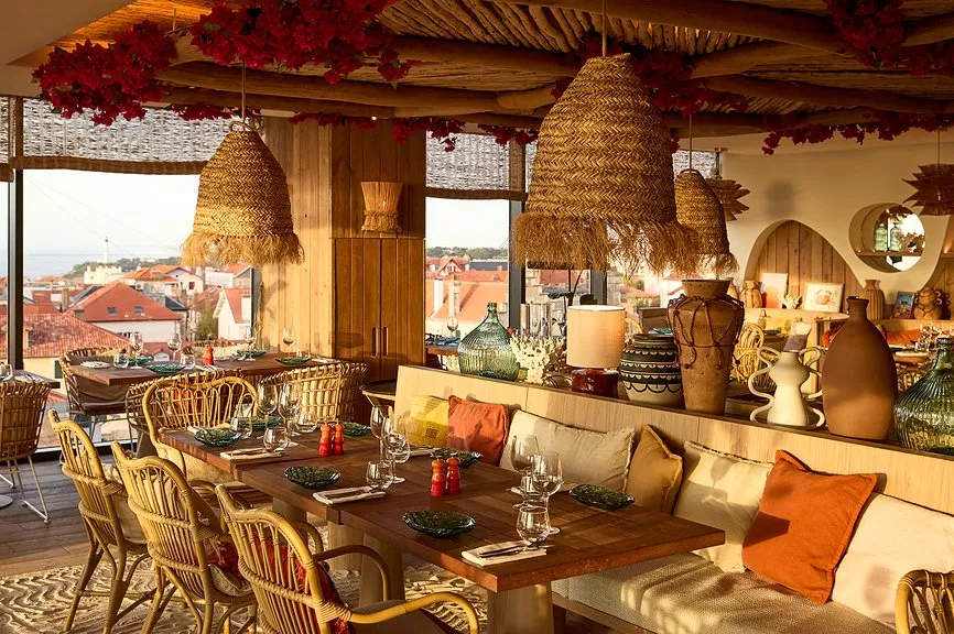 Intérieur chaleureux d'un restaurant avec plafond en bois brut, bouganvilliers rouges, suspensions en osier, tables en teck et vue dorée sur les toits de Biarritz.