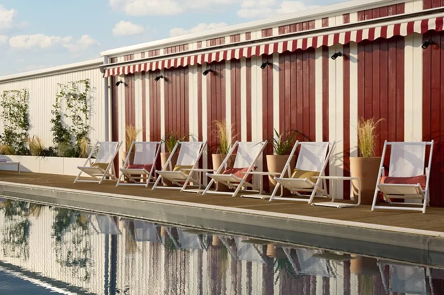 Rangée de transats blancs au bord d'une piscine sur rooftop, devant une façade à rayures bordeaux et blanc avec auvent assorti.