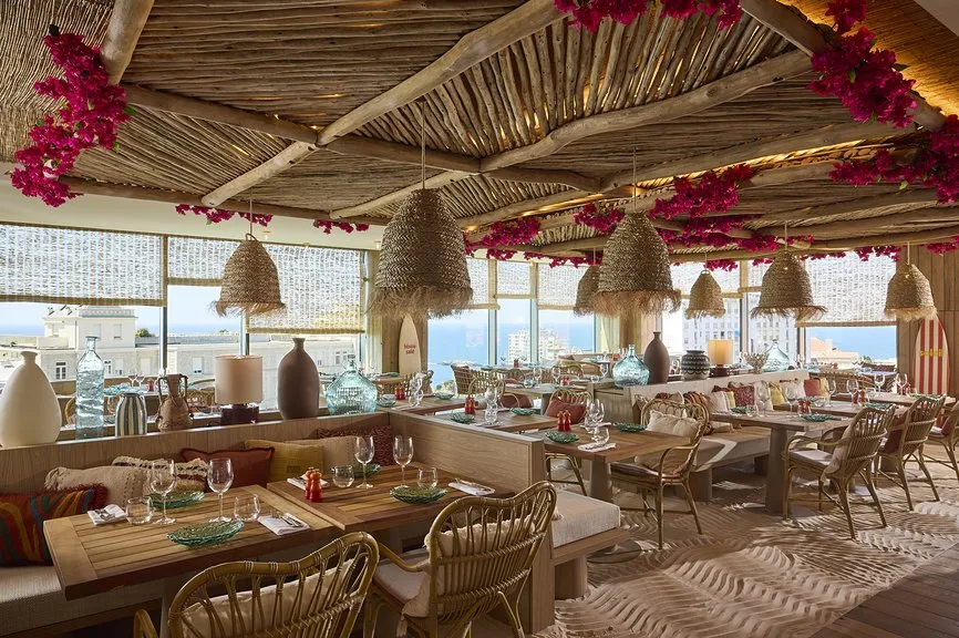 Salle de restaurant estivale avec plafond en bois brut orné de bouganvilliers fuchsia, suspensions en osier, tables en teck et vue sur l'océan.