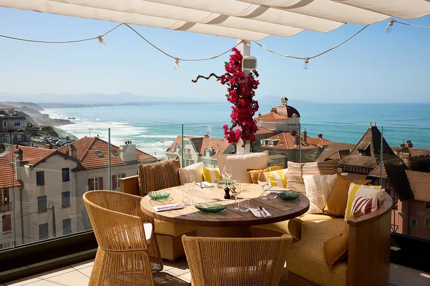 Table ronde dressée sur rooftop avec fauteuils en rotin et bouganvillier fleuri, vue panoramique sur les toits de Biarritz, la côte et l'océan.