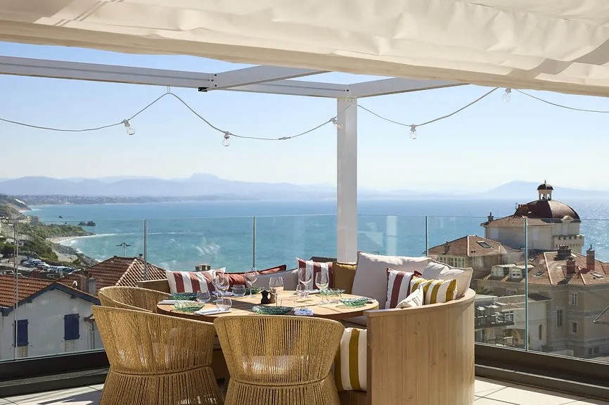 Table dressée en rotin sur rooftop terrasse avec pergola et guirlande lumineuse, offrant une vue panoramique sur les toits de Biarritz et l'océan.