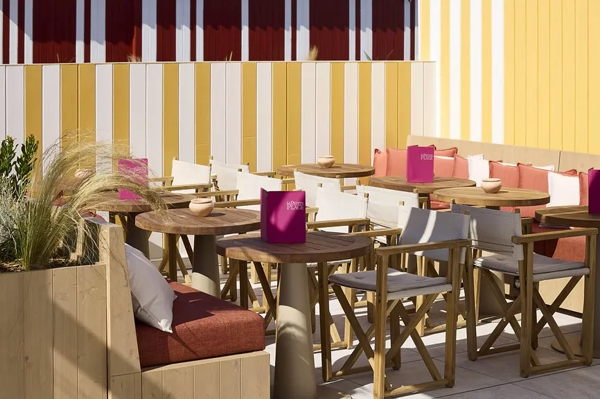 Terrasse de restaurant ensoleillée avec tables et chaises en teck, banquettes colorées et façade à rayures jaunes et bordeaux.