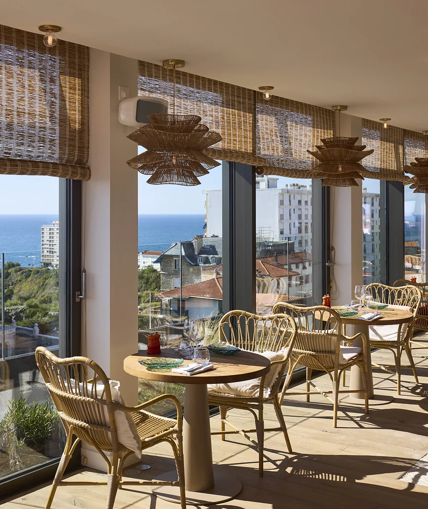 Salle de restaurant lumineuse avec tables et chaises en rotin, suspensions en fibres naturelles et baies vitrées panoramiques vue sur Biarritz et l'océan.