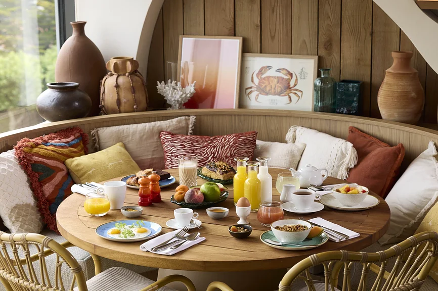 Petit-déjeuner généreux dressé sur table ronde en bois dans une banquette arrondie en bois, entourée de coussins colorés, vases en terre cuite et illustrations marines.