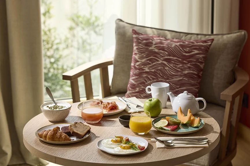 Petit-déjeuner complet servi en chambre sur table ronde : viennoiseries, œufs, charcuterie, fruits frais, jus et théière, devant une fenêtre lumineuse.