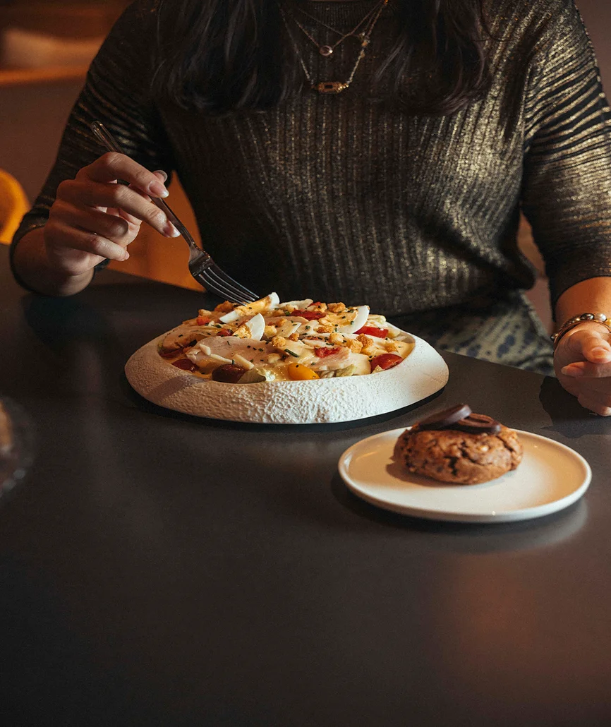 Une femme dégustant une salade niçoise dressée sur assiette texturée, avec un cookie au chocolat en accompagnement, dans une ambiance de restaurant tamisée.