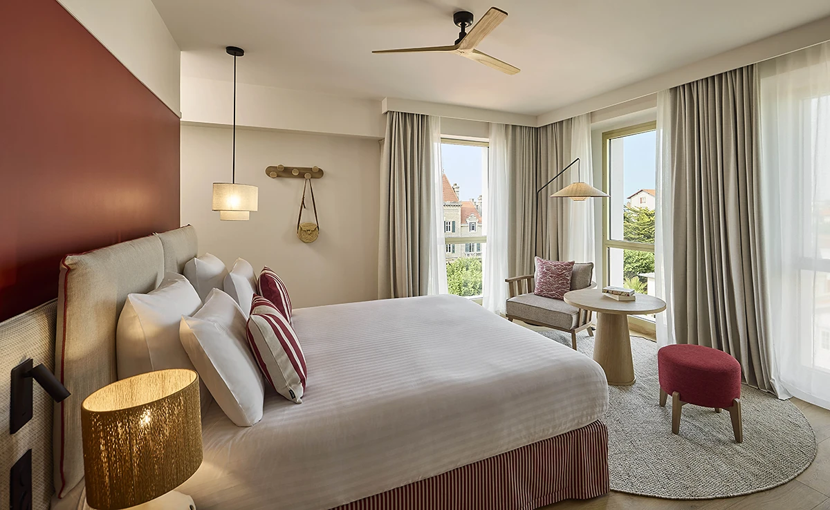 Chambre d'hôtel spacieuse avec grand lit, ventilateur au plafond, mur terracotta et vue sur les toits de Biarritz