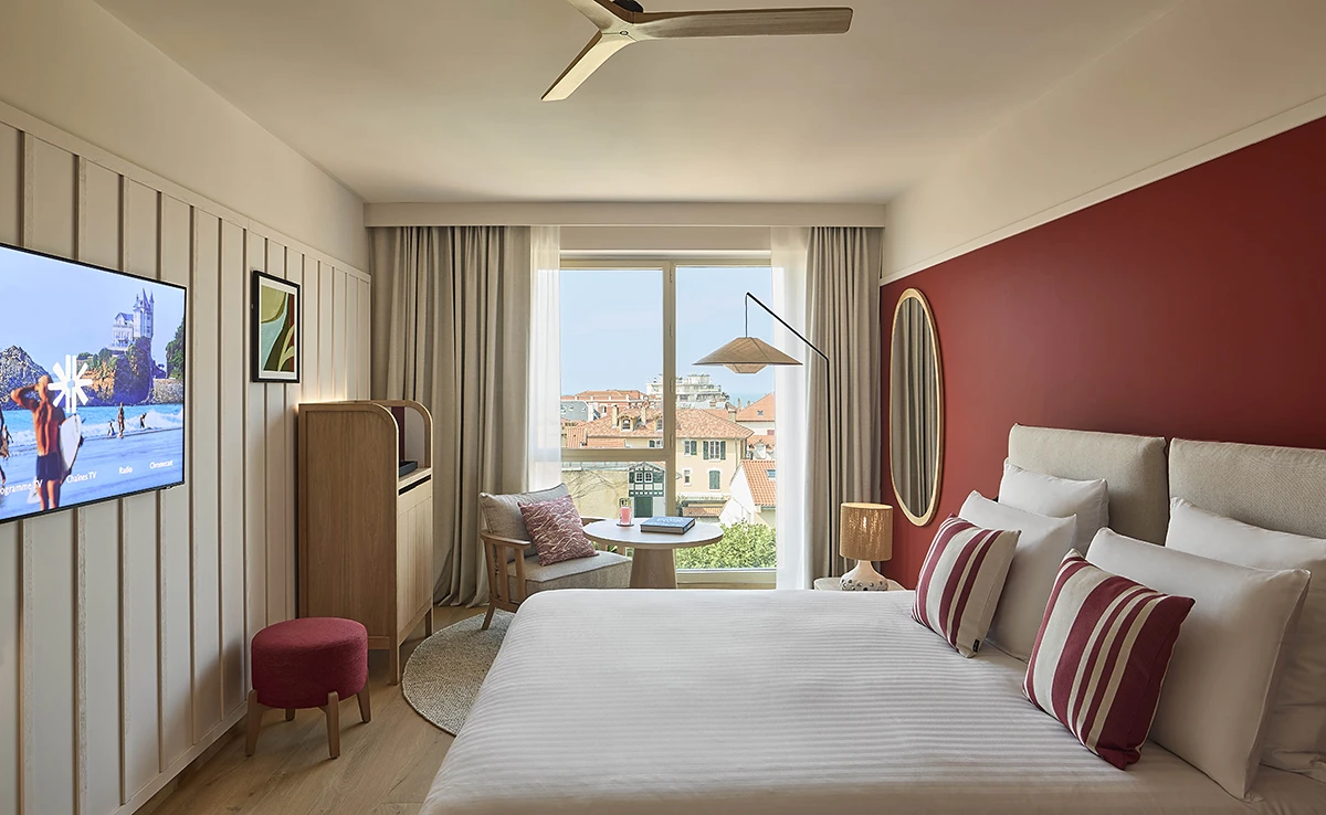 Chambre d'hôtel avec grand lit, télévision murale, mur terracotta, miroir ovale et vue sur les toits de Biarritz
