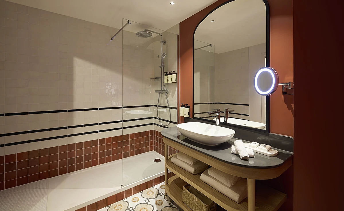 Salle de bain d'hôtel avec vasque posée, baignoire et douche carrelées terracotta et blanc, miroir arrondi rétroéclairé