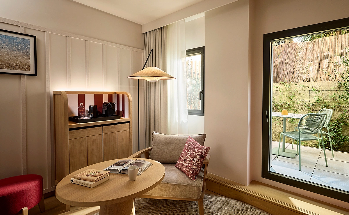Hotel room lounge area with an armchair, wooden coffee table, and direct access to a lush private terrace.