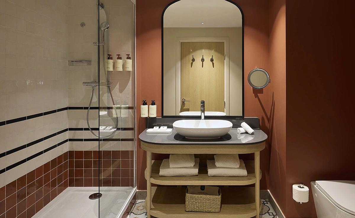 Salle de bain d'hôtel avec vasque sur meuble en bois, douche à l'italienne carrelée terracotta et blanc, murs bordeaux