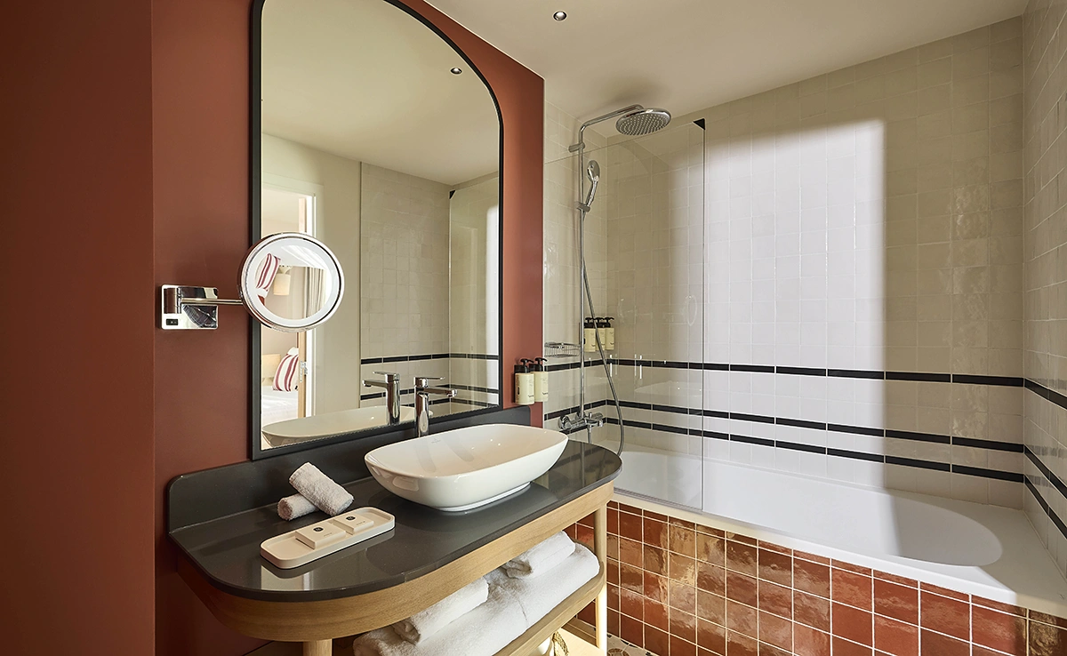 Salle de bain d'hôtel élégante avec vasque posée sur meuble en bois, baignoire et douche, carrelage terracotta et blanc aux accents noirs