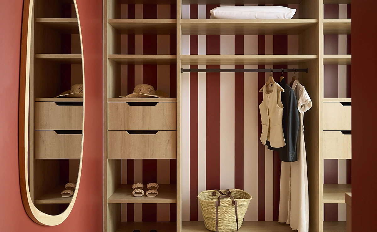 Dressing ouvert en bois clair avec fond rayé bordeaux, miroir ovale et penderie dans une chambre de l'hôtel Le Talaia Mgallery Biarritz