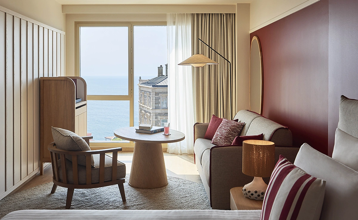 Coin salon d'une chambre de l'hôtel Le Talaia Mgallery Biarritz avec canapé, fauteuil en bois et vue sur l'océan