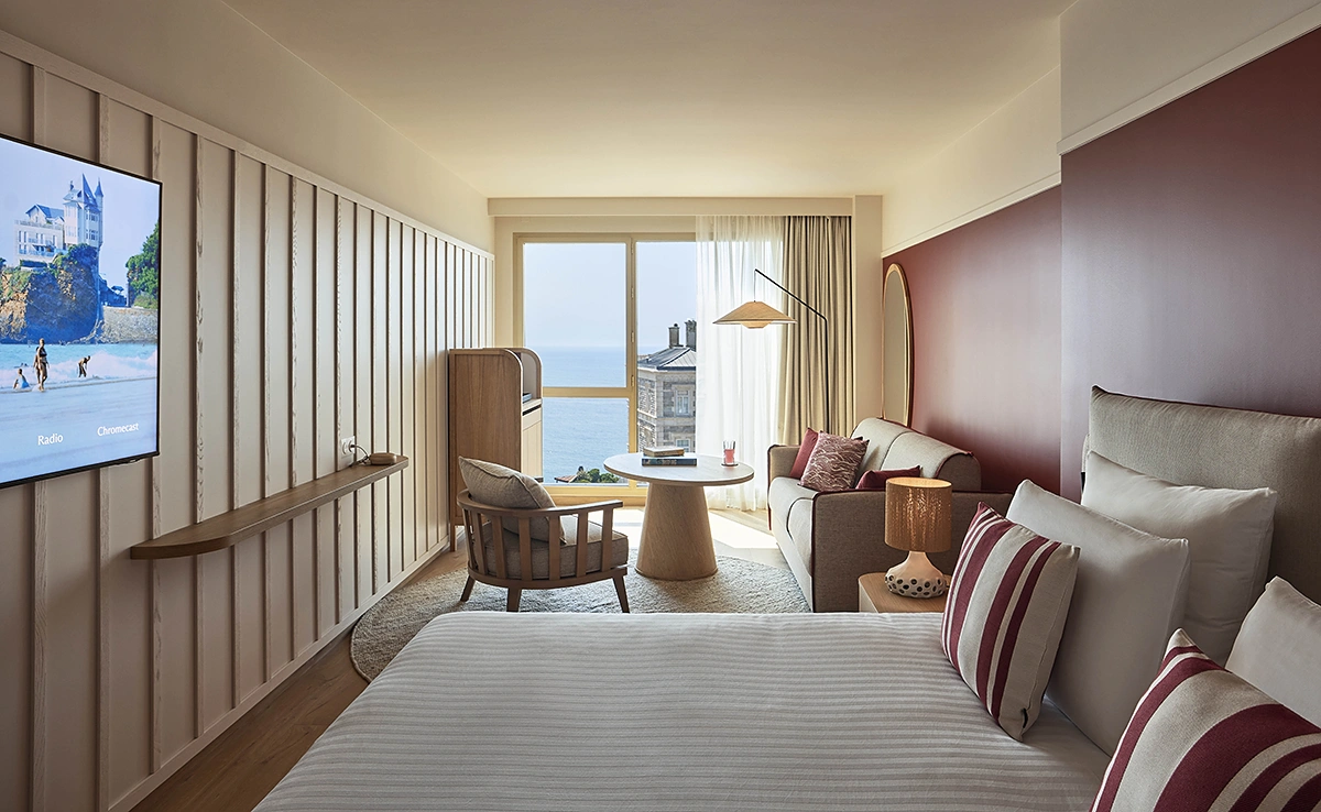 Chambre avec vue océan de l'hôtel Le Talaia Mgallery Biarritz avec lit, coin salon, télévision murale et mur bordeaux