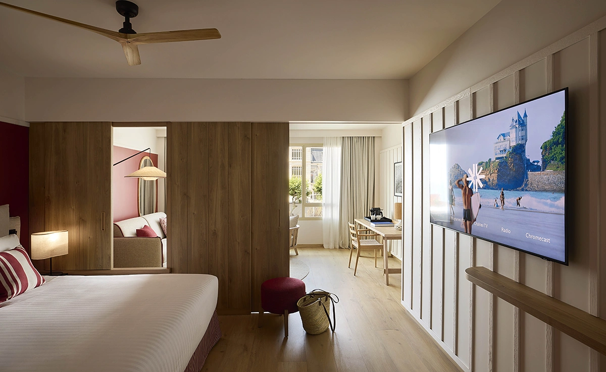 Vue d'ensemble d'une chambre de l'hôtel Le Talaia Mgallery Biarritz avec lit, télévision murale affichant un paysage de Biarritz, bureau et ventilateur de plafond