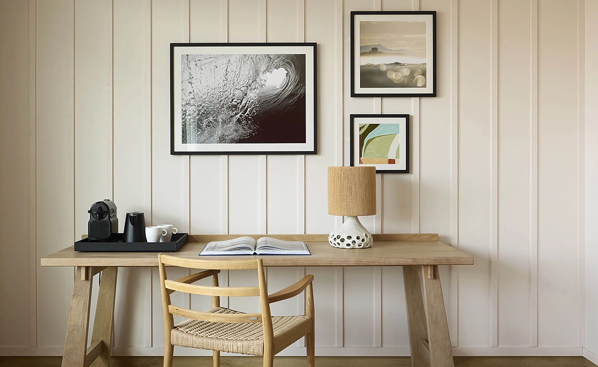 Bureau en bois clair d'une chambre de l'hôtel Le Talaia Mgallery Biarritz avec machine Nespresso, lampe en raphia et galerie de photos surf au mur