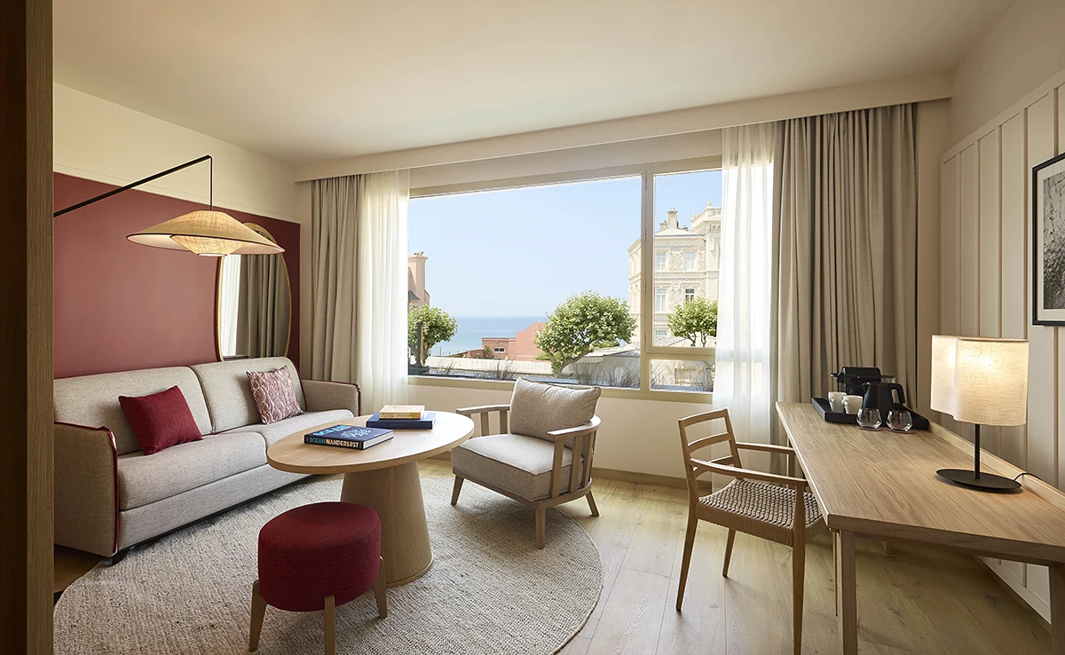 Salon lumineux d'une suite de l'hôtel Le Talaia Mgallery Biarritz avec canapé beige, accents bordeaux, bureau en bois et vue sur l'océan