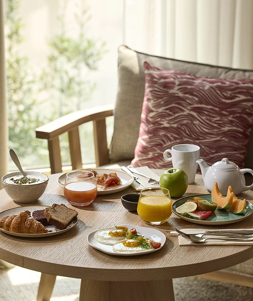 Petit-déjeuner complet servi sur table ronde en bois clair : viennoiseries, œufs, charcuterie, fruits frais, jus et théière, dans un coin lumineux.