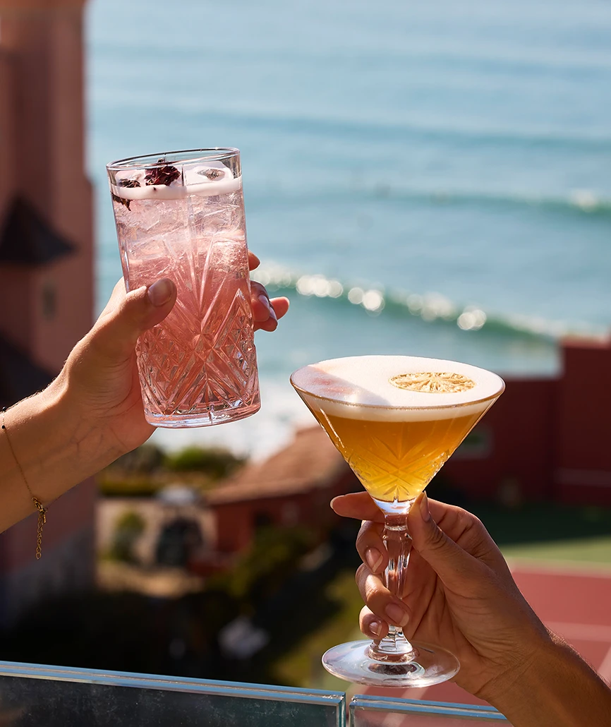 Deux mains portant un toast avec un highball rose et un martini doré à l'écume, en terrasse face à l'océan de Biarritz.