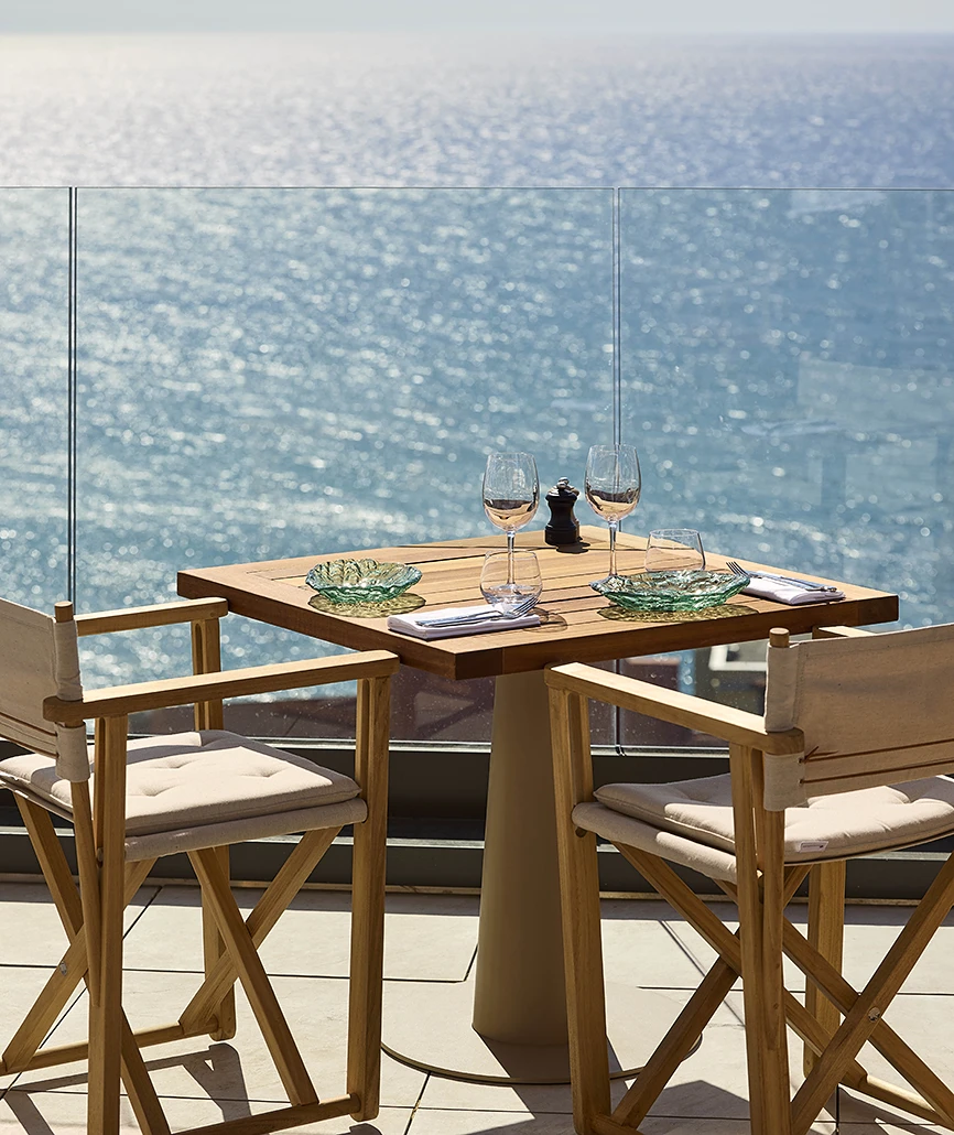 Table dressée pour deux en terrasse avec verres à vin et coupelles en verre soufflé, face à l'océan scintillant derrière une balustrade en verre.