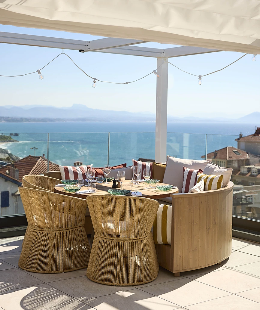 Salon de rooftop avec fauteuils et canapé en rotin tressé autour d'une table dressée, sous pergola avec vue panoramique sur l'océan et les montagnes.