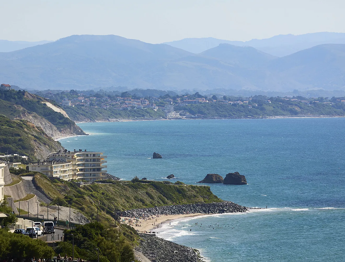 Vue panoramique sur la côte basque avec falaises, plages de surf, rochers émergents et les Pyrénées en arrière-plan.