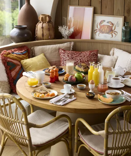 Table ronde en bois dressée pour le petit-déjeuner avec fruits, jus et viennoiseries, entourée de chaises en rotin au restaurant de l'hôtel Le Talaia Mgallery Biarritz