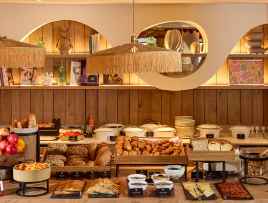 Buffet de petit-déjeuner de l'hôtel Le Talaia Mgallery Biarritz avec viennoiseries, pains et céréales sous un luminaire en raphia à franges