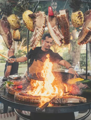 Black-clad chef grilling food on a large, circular outdoor barbecue with open flames, with cuts of meat and pineapples hanging above the fire.