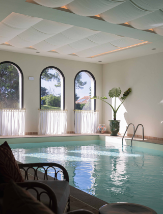 An indoor hotel or spa pool featuring a white undulating fabric ceiling, large arched windows draped with white curtains, a tropical plant, and rattan chairs by the poolside.