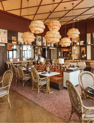 Elegant dining room in warm terracotta and wood tones, featuring golden rattan chairs, camel leather banquettes, large woven wicker pendant lights, and walls adorned with frames, creating a chic brasserie ambiance.