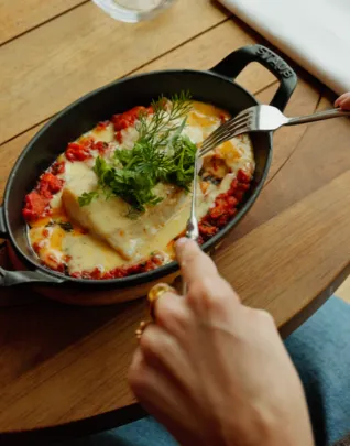 Main tenant une fourchette au-dessus d'un plat cuisiné servi en cocotte noire ovale, garni de fromage fondu, sauce tomate et herbes fraîches, posé sur une table en bois