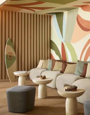 Espace lounge design et coloré avec un canapé arrondi blanc, des tables basses en forme de champignon, des poufs gris, une planche de surf vintage appuyée contre un mur à lamelles en bois, devant une fresque murale aux formes organiques dans des tons terracotta, vert sauge et bleu pastel