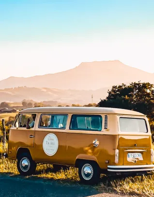 Volkswagen Combi vintage de couleur ocre jaune stationné dans un paysage de campagne au coucher du soleil, avec une montagne brumeuse en arrière-plan et une végétation dorée, dans une atmosphère chaleureuse et nostalgique