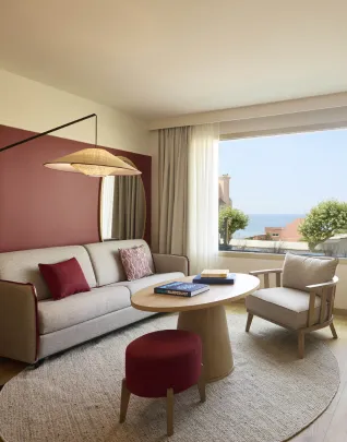 Sala de estar de una elegante habitación de hotel con un sofá beige, una mesa de centro redonda de madera, un sillón y un puf burdeos, un gran ventanal con vistas al mar, y una decoración en tonos cálidos rosa y crema