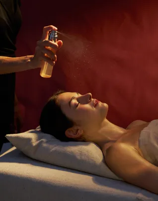 Vue en contre-plongée d'une femme allongée sur une table de soin, visage détendu tourné vers le brouillard d'un spray aromatique vaporisé par un thérapeute, sur fond de mur bordeaux baigné d'une lumière chaude et dramatique