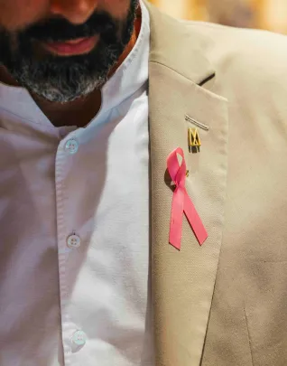 Gros plan sur le revers d'une veste beige portée par un homme barbu, avec un ruban rose symbole de la lutte contre le cancer du sein épinglé à la poche